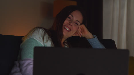 Smiling-Woman-Sitting-On-Sofa-At-Home-At-Night-Streaming-Or-Watching-Movie-Or-Show-On-Laptop-1